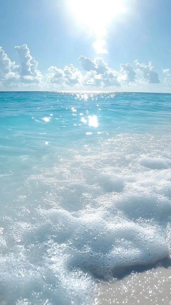 The image shows a beautiful beach scene with clear blue water and white foamy waves gently lapping at the shore. The sun shines brightly in the sky, creating a sparkling effect on the water. The sky is a soft blue with fluffy white clouds scattered across it. The scene is tranquil and inviting, evoking a sense of peace and relaxation.