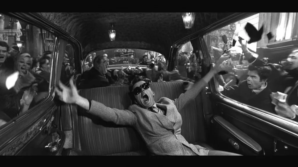 A man in a suit and sunglasses sits in the back of a car, his arms outstretched, screaming in a black and white photo.  A crowd of people can be seen outside the car's windows, some reaching towards the man inside. The scene is chaotic and suggests a moment of crisis.