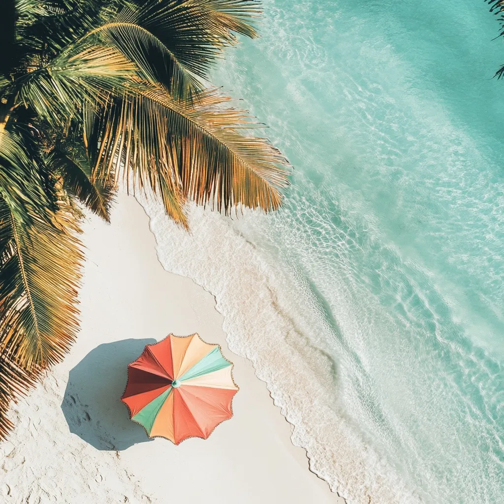 An aerial view of a tropical beach with crystal clear turquoise water. White sand and foamy waves lap against the shore, beneath the lush green fronds of a palm tree. A colorful beach umbrella stands on the sand, creating a perfect spot for relaxation and enjoying the summer sunshine.