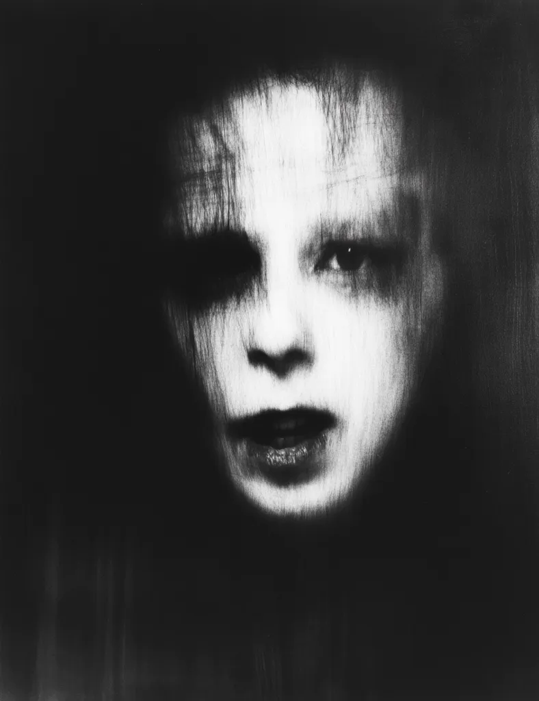 The image is a black and white portrait of a person's face. The face is partially obscured by darkness, with only the eyes, nose, and mouth visible. The eyes are wide open and staring, while the mouth is slightly open. The face has a ghostly and eerie appearance, as if it were a spirit or a ghost. The image is likely meant to evoke feelings of fear or unease.
