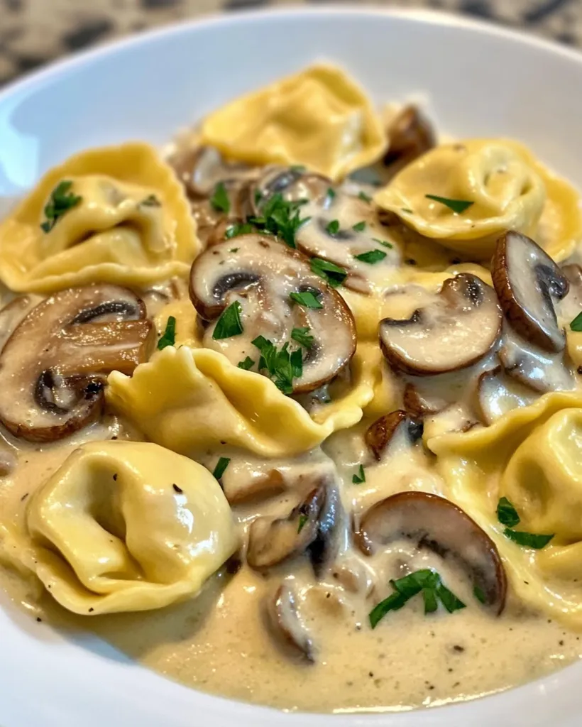 A white bowl filled with a creamy mushroom sauce and tortellini pasta. The sauce is thick and creamy, and the mushrooms are cooked to perfection. The tortellini pasta is soft and chewy, and the dish is garnished with fresh parsley. The dish is visually appealing and looks delicious.