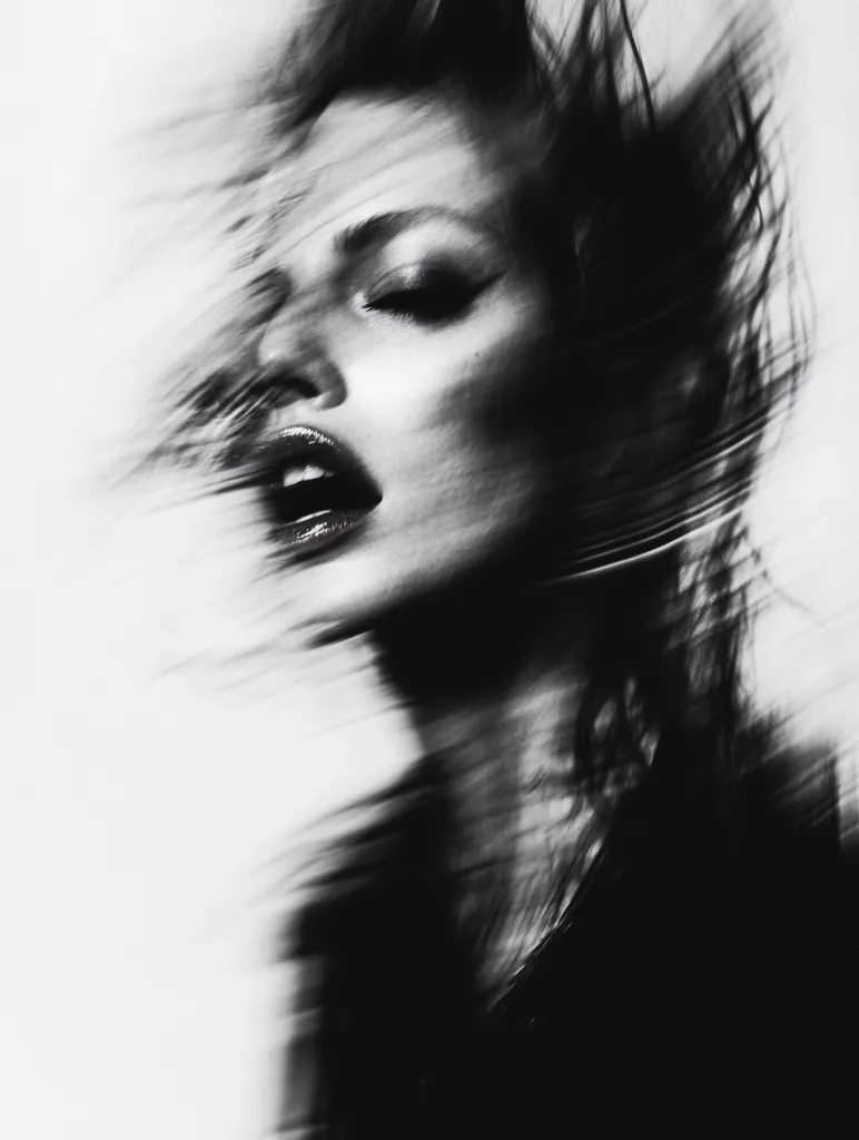 A black and white portrait of a woman with her hair flowing dramatically. Her face is partially obscured by the movement of her hair, creating a sense of mystery and intrigue. The image is shot in a high-contrast style, emphasizing the sharp lines and textures of her features. The overall effect is one of raw beauty and captivating sensuality.