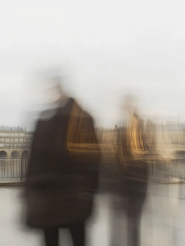 Two figures are blurred in motion, walking in front of a city scene with buildings and a railing. The figures are out of focus, creating an abstract and mysterious composition. The image captures a sense of movement and anonymity in the city. The blurred background suggests a bustling urban environment.