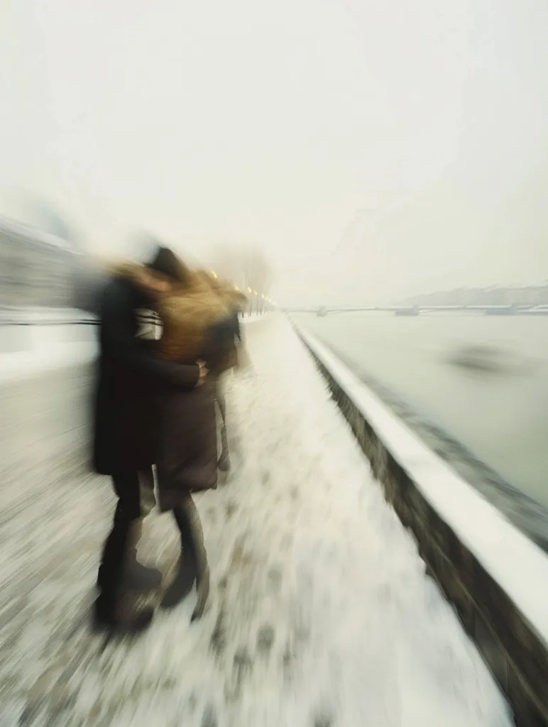 A couple walks along a snowy walkway, their figures blurred in motion. The scene is bathed in a hazy, overcast light, with a body of water visible in the distance. The photo has a dreamlike quality, emphasizing the fleeting nature of the moment.