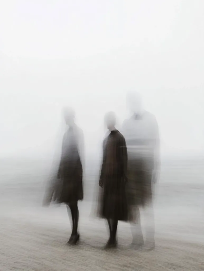The image depicts three blurred figures standing in a foggy landscape. The figures are out of focus, creating an ethereal and dreamlike atmosphere.  They are dressed in dark clothing, and their forms are barely discernible against the white background. The overall impression is one of mystery and ambiguity.
