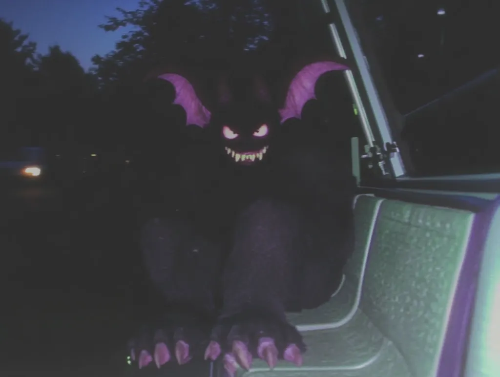 A dark, shadowy figure with glowing purple eyes and ears sits on a green seat in the back of a vehicle. The figure has sharp claws and a menacing grin. The scene is set at night, with faint lights in the distance and trees surrounding the vehicle. The image evokes a sense of unease and mystery.