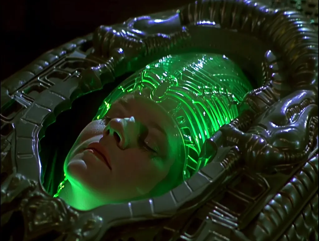 A woman's face is visible, lying in a metallic and biomechanical device.  The device is green and glowing.  The woman is unconscious, with her eyes closed and her mouth slightly open.  The device appears to be some kind of alien technology.  The woman's face is pale and her lips are slightly parted.