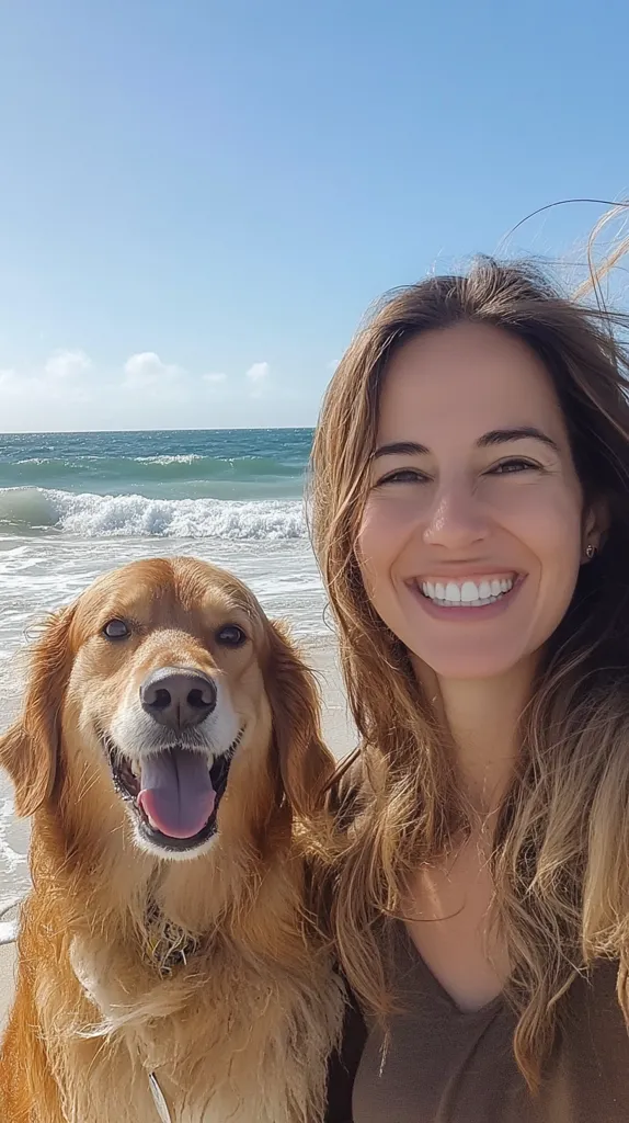 A young woman with long brown hair smiles at the camera while standing on a beach with a golden retriever. The dog is looking at the camera with its tongue sticking out. The ocean waves are crashing in the background.  The sky is blue and the day is sunny.