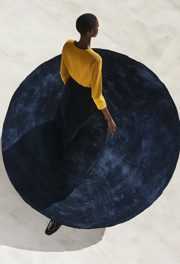 A woman with short dark hair is wearing a yellow sweater and a long, flowing blue skirt. She is standing on a white surface with her skirt forming a circle around her. The skirt has a textured, woven appearance, and the woman's silhouette is partially visible through the fabric. The image captures a minimalist, fashion-focused moment.