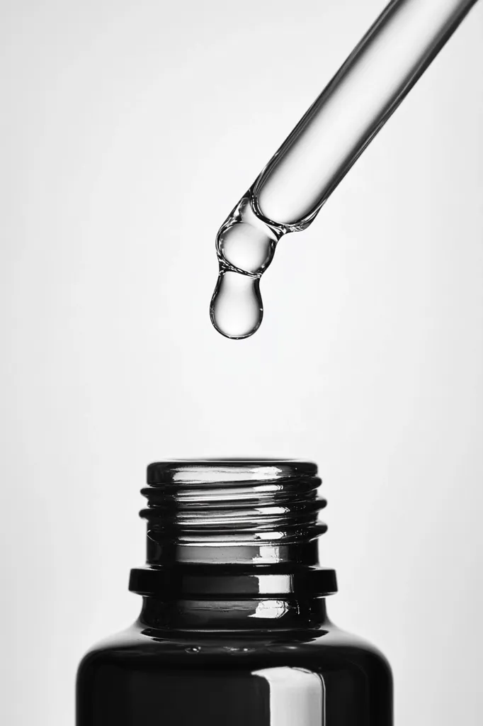 A glass dropper is positioned above an amber glass bottle, about to dispense a drop of liquid. The dropper is clear and the liquid inside is translucent, forming a teardrop at the tip. The image is minimalist and focuses on the precision of the moment, emphasizing the act of dispensing the liquid. The background is a simple white, allowing the focus to remain on the dropper and bottle.