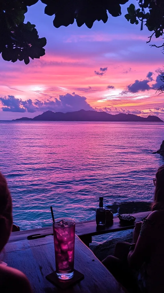 Here's a description of the image:

The photograph captures a breathtaking sunset over a tranquil ocean. The sky is ablaze with vibrant hues of pink, purple, and orange, reflecting beautifully on the water's surface.  A silhouette of a distant island adds depth to the scene.  In the foreground, a person sits at an outdoor table, enjoying a purple cocktail as the sun dips below the horizon.  The overall mood is serene and romantic, enhanced by the dark silhouettes of overhanging tree branches framing the sunset. The scene is peaceful and idyllic, suggesting a tropical location.
