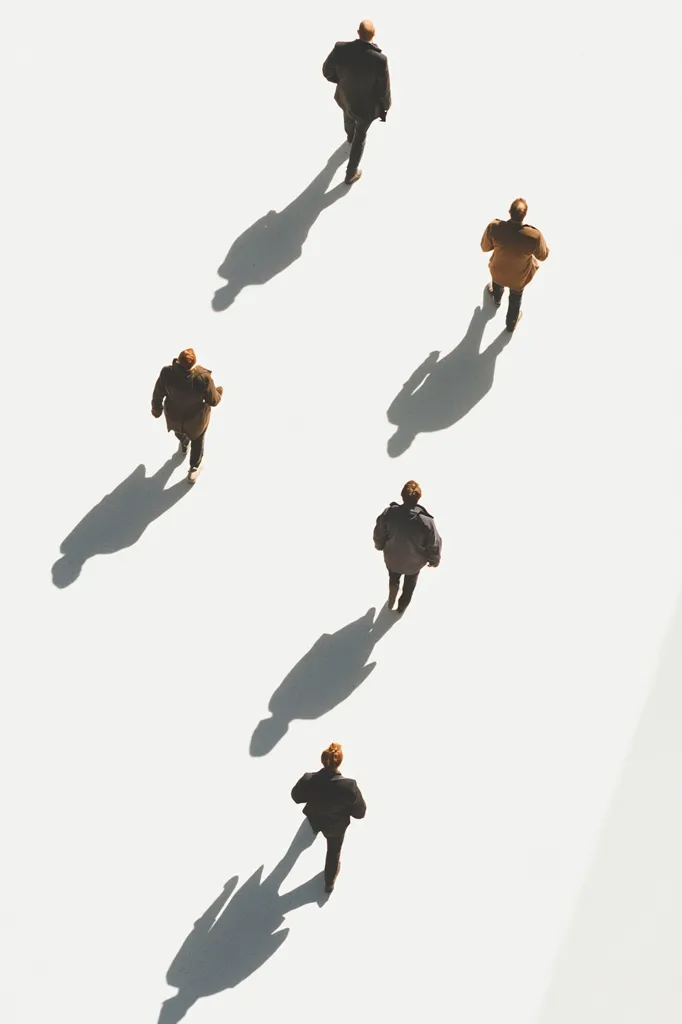 Here's a description of the image:

The image shows five individuals, dressed in business casual attire, walking in a staggered, diagonal line across a bright white surface.  Each person casts a distinct shadow that stretches behind them, indicating a strong overhead light source. The arrangement suggests a sense of movement and individual progress, yet within a shared trajectory. The overall aesthetic is minimalist and clean, emphasizing the figures and their shadows against the stark white background.