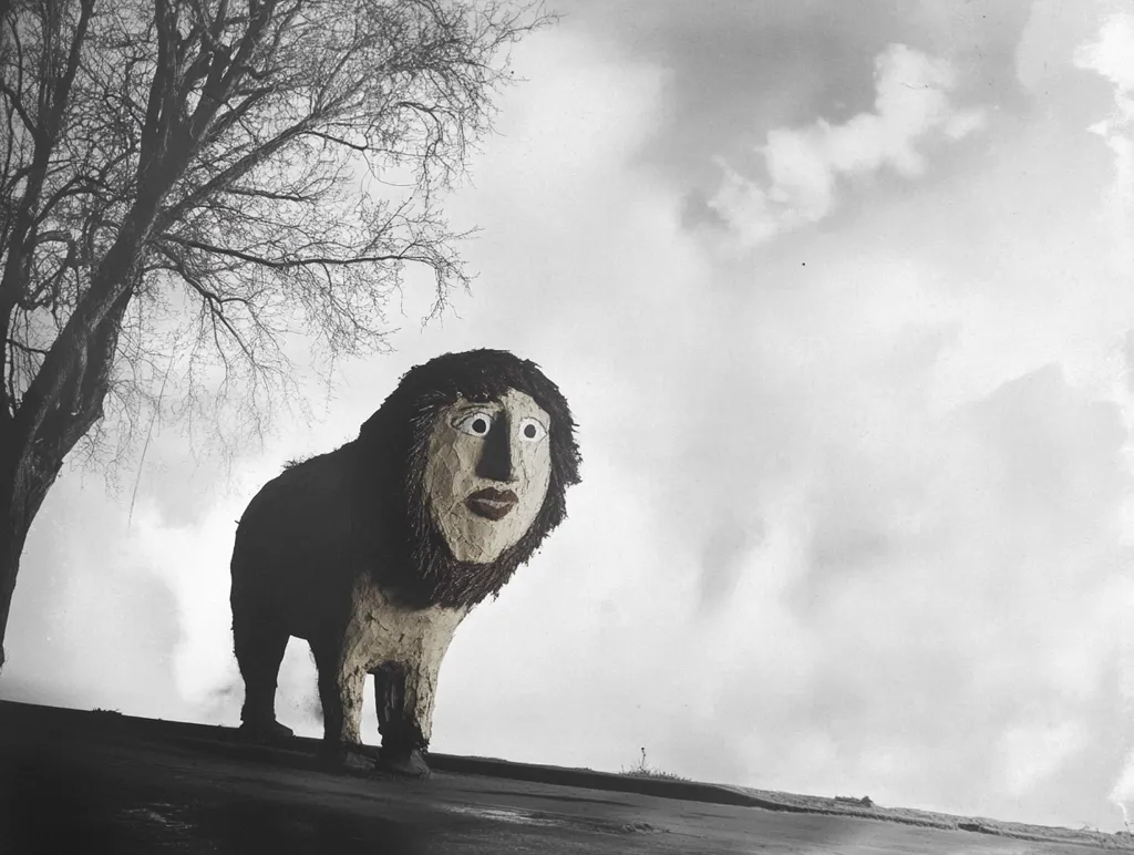 Here's a description of the image:

The monochromatic photograph depicts a peculiar, lion-like figure constructed from what appears to be sculpted materials.  Its most striking feature is a human-like face, with expressive eyes and lips, affixed to the creature's head. The lion stands on a dark, seemingly paved surface, beneath a stark, leafless tree and a pale, cloudy sky.  The overall mood is surreal and slightly unsettling, the contrast between the creature's features and its animalistic form creating a sense of unease. The background is muted, drawing attention to the strangely compelling figure in the foreground.