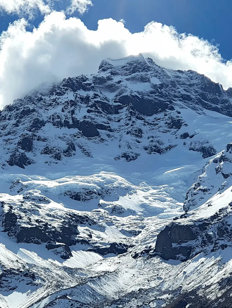A majestic snow-capped mountain peak dominates the image, partially veiled by fluffy white clouds under a brilliant blue sky.  The mountain's rugged, rocky terrain is heavily covered in snow and ice, with visible glaciers carving their way down the slopes.  The contrast between the bright white snow, dark rock faces, and vibrant blue sky creates a dramatic and awe-inspiring scene.  The lower slopes reveal a textured landscape of snow-dusted rocks and ice formations.