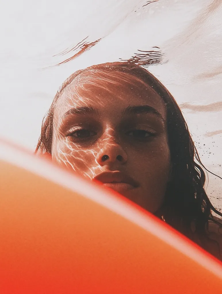 Here's a description of the image:

The close-up shot shows a person's face partially submerged in water, their gaze directed towards the viewer.  Sunlight refracts through the water, creating shimmering patterns across their skin.  Only the upper half of their face is visible, framed by dark, wet hair. A vibrant orange object, possibly an inflatable, obscures the lower half of the face and the bottom of the frame. The overall tone is warm and slightly hazy, evoking a summery, underwater ambiance. The image is aesthetically pleasing, with a focus on texture and light.