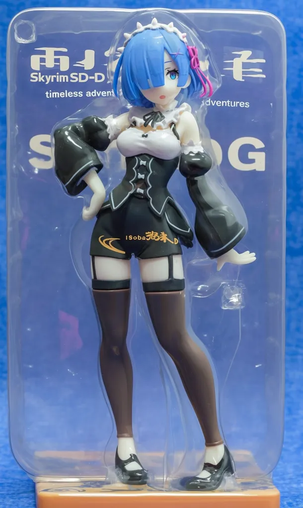 This is a close-up shot of a Rem figurine from the anime Re:Zero.  The figurine is displayed in its clear plastic packaging, which shows the character in a maid outfit with dark stockings and shoes. The packaging features text in both English and Japanese, including "Skyrim SD-D timeless adventures," hinting at a collaboration or themed release. The figurine is posed with a hand on her hip, showcasing details like her signature blue hair and dress. The base is a muted orange.