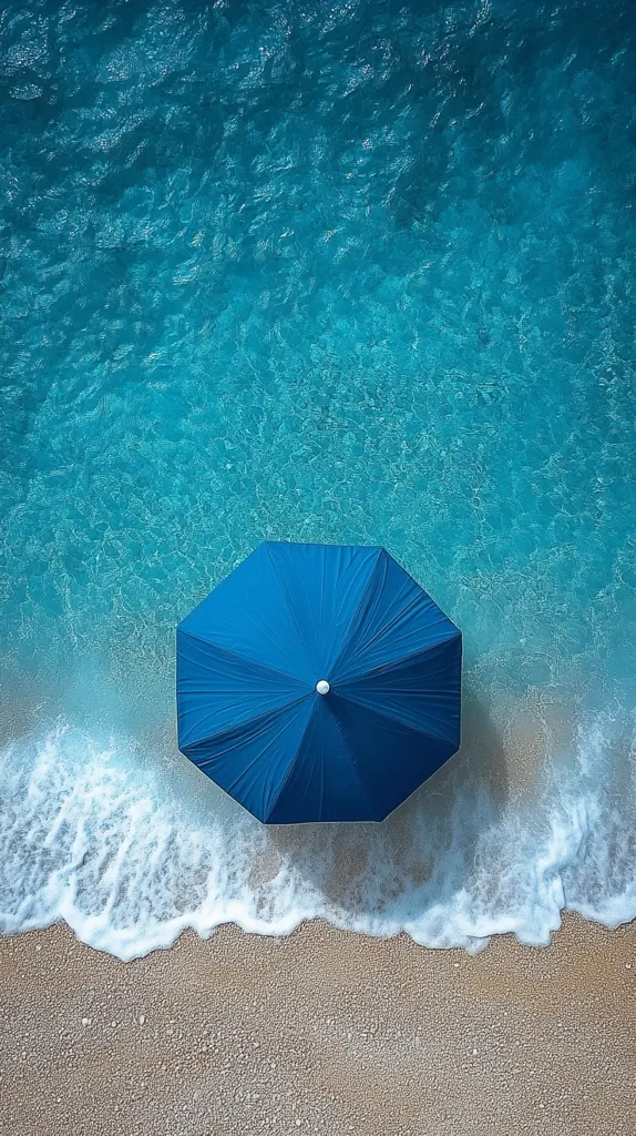 Here's a description of the image:

An overhead shot captures a vibrant scene of a beach.  Crystal-clear turquoise water laps gently at a sandy shore, where a single, closed, dark-blue beach umbrella sits.  White foam from the waves creates a delicate line between the water and the sand. The overall aesthetic is one of serene tranquility and summer vacation. The colors are bright and contrasting, with the blue of the water and umbrella standing out against the lighter beige of the sand.