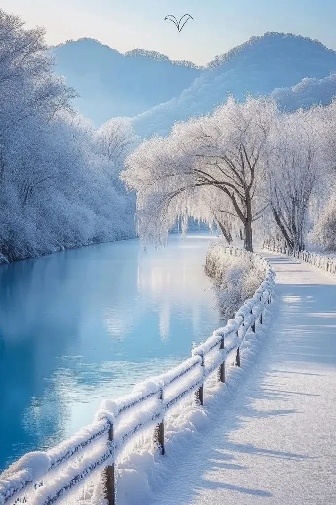A serene winter scene unfolds along a snow-covered path beside a tranquil river.  Frost-laden trees, particularly a striking weeping willow, line the riverbank, their branches heavy with glistening ice crystals. A dark wooden fence, also dusted with snow, guides the way, leading towards a backdrop of softly hued, snow-dusted mountains.  The river's surface reflects the pale blue sky, creating a harmonious and idyllic winter wonderland.  A small bird is visible in the upper portion of the image.