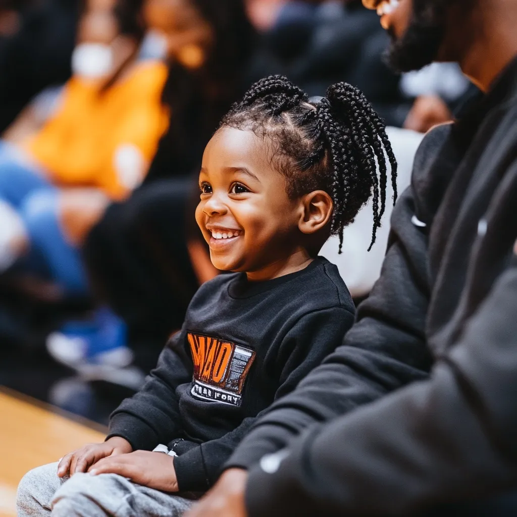 Here's a description of the image:

Close-up view of a young Black child with adorable braided pigtails, seated and beaming with a radiant smile. The child is wearing a dark-colored sweatshirt with an orange graphic design.  They are sitting next to an adult, possibly a parent, whose dark clothing partially obscures the background.  The setting appears to be an indoor arena, suggested by the wooden floor visible at the bottom of the image and the blurred figures in the background, possibly a crowd of people. The overall atmosphere is warm and intimate, capturing a tender moment.