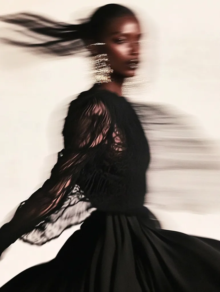 Here's a description of the image:

The photograph showcases a dark-skinned woman in motion.  Her black, sheer-sleeved top and flowing black skirt are blurred, suggesting rapid movement.  Her long dark hair streams behind her, adding to the sense of dynamism.  She wears long, sparkling gold earrings. The overall aesthetic is elegant and slightly mysterious, with the motion blur creating a dreamy, ethereal effect against a neutral background.  The focus is on the woman's form and the feeling of movement, rather than sharp detail.