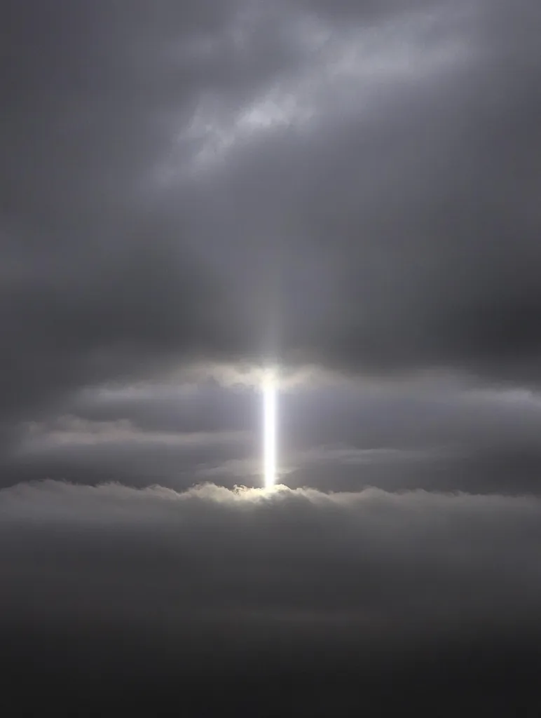 A dramatic image depicts a slender beam of bright light piercing through a dense layer of dark, ominous clouds.  The light originates from above, illuminating a small portion of the lower cloud cover, creating a stark contrast between the shadowy atmosphere and the radiant beam. The overall mood is one of awe and mystery, suggesting a powerful, ethereal presence breaking through darkness.