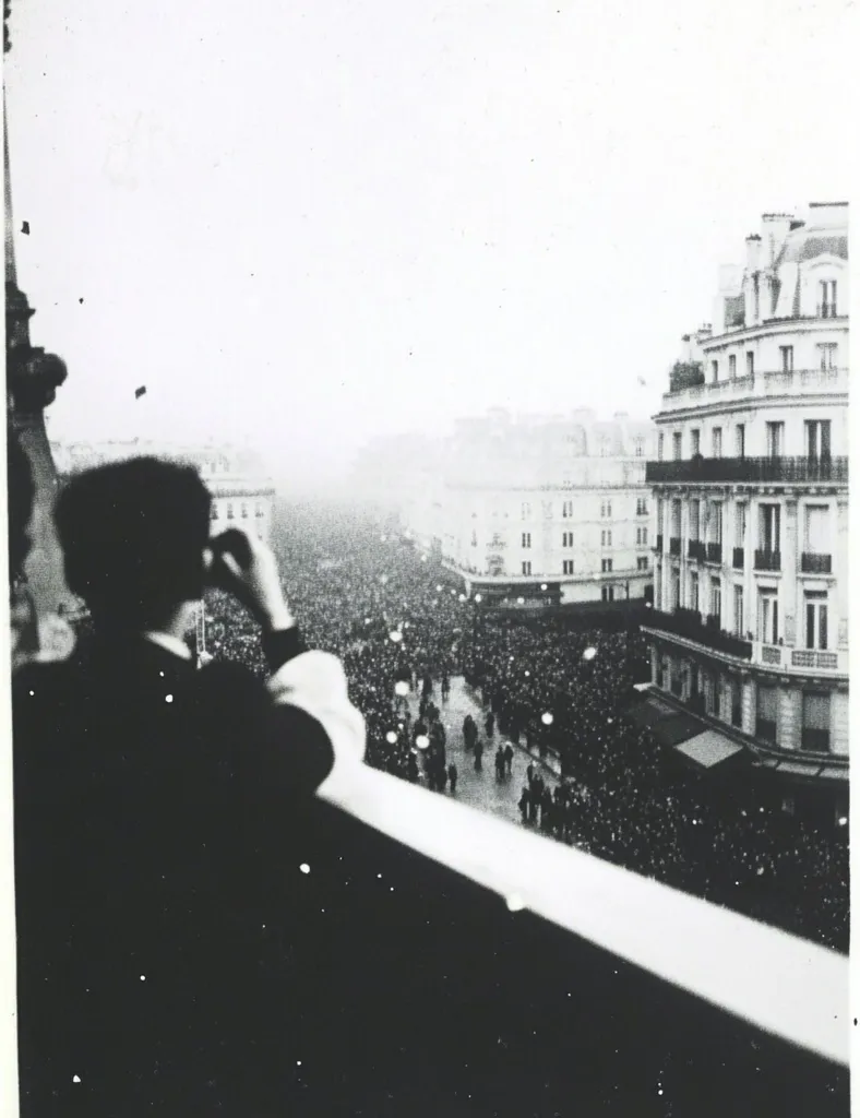Here is a description of the image:

A black and white photograph captures a vast crowd surging through a Parisian street, viewed from an elevated vantage point. The observer, partially obscured, is silhouetted in the foreground, their hand raised to their face, possibly shading their eyes or shielding themselves from the elements.  The street scene stretches into the distance, eventually fading into a hazy background. Elegant Parisian buildings line the street, their architectural details visible despite the distance and the overall hazy atmosphere.  The image suggests a significant public event or procession, possibly a parade or celebration, due to the size of the crowd. The photograph has a grainy, slightly washed-out quality characteristic of older prints.