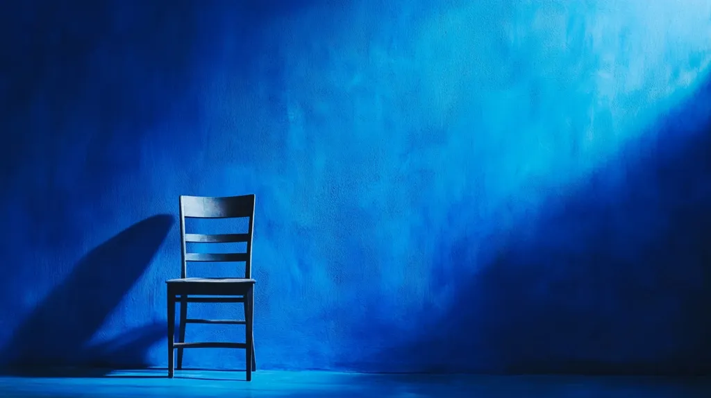 A single, dark-colored wooden chair sits against a vibrant blue wall.  The wall has a textured, uneven finish, and a dramatic light source casts a long shadow of the chair to its left. The overall mood is somber and contemplative, with the stark contrast between the chair and the background creating a striking visual effect. The image is minimalist in composition, focusing attention on the solitary chair in a spacious setting.
