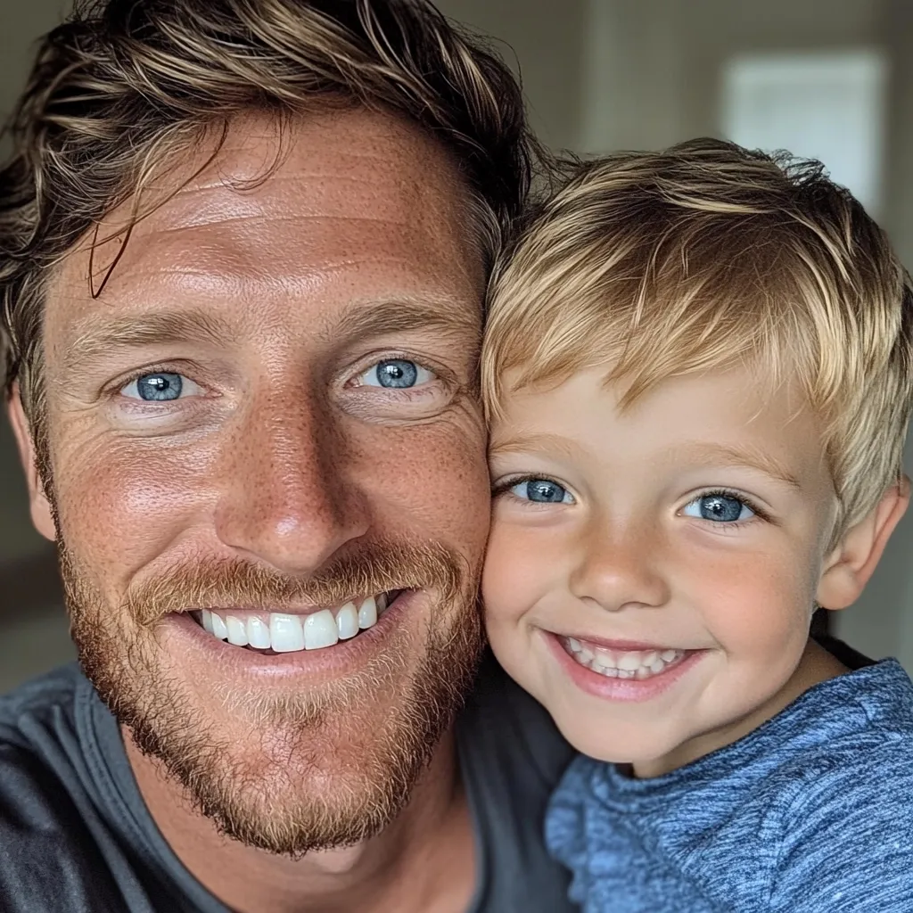 Here's a description of the image:

Close-up selfie of a man and a young boy. The man has light brown, slightly wavy hair, blue eyes, and a reddish-brown beard. He's smiling broadly, showcasing a bright, white smile. The boy, with light blonde hair and also blue eyes, is nestled against the man's shoulder, mirroring his smile.  Both have fair complexions. The background is blurred, suggesting an indoor setting. The overall impression is one of warmth, affection, and a close father-son bond.