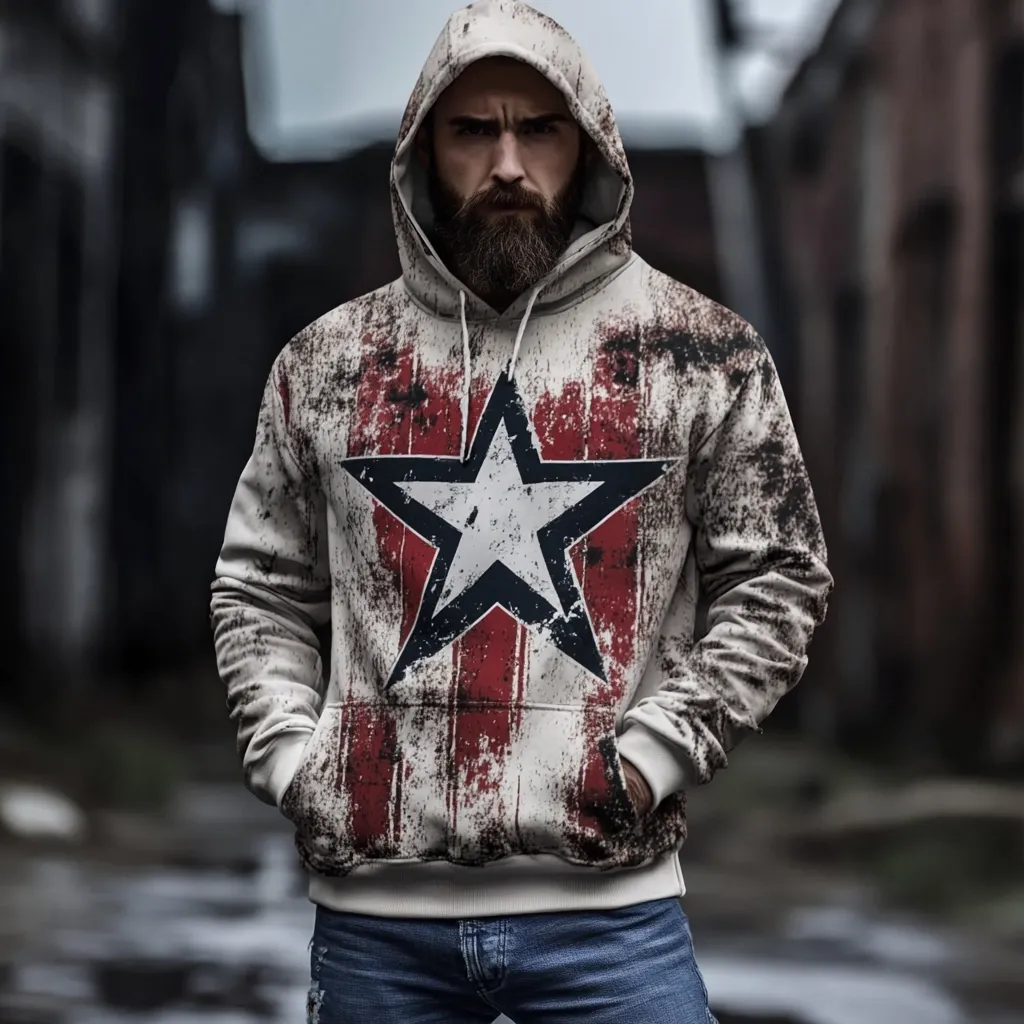 A bearded man with a serious expression stands outdoors wearing a cream-colored hoodie. The hoodie features a large, distressed red and white star graphic reminiscent of an American flag. The style is casual, with a worn or vintage look.  He has his hands in his pockets, and the background is blurred, suggesting an urban setting. The overall image conveys a blend of ruggedness and patriotism.