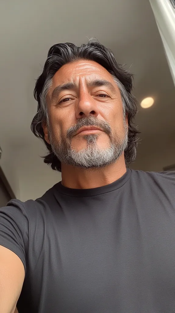 Close-up selfie of a man with salt-and-pepper hair and a short, well-groomed beard. He's wearing a dark gray crew-neck t-shirt.  His expression is serious, almost pensive. The background is blurred, suggesting an indoor setting. The lighting is soft and natural. The overall image conveys a sense of calm and maturity.