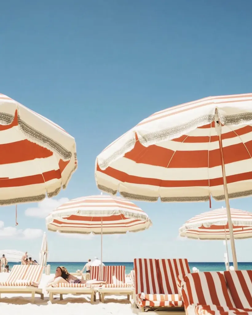 Red and white striped beach umbrellas shade rows of matching lounge chairs on a pristine, sandy beach. A woman relaxes on one of the chairs, enjoying the sunny day.  The clear blue sky provides a vibrant backdrop to this idyllic beach scene.  Other beachgoers are visible in the distance.