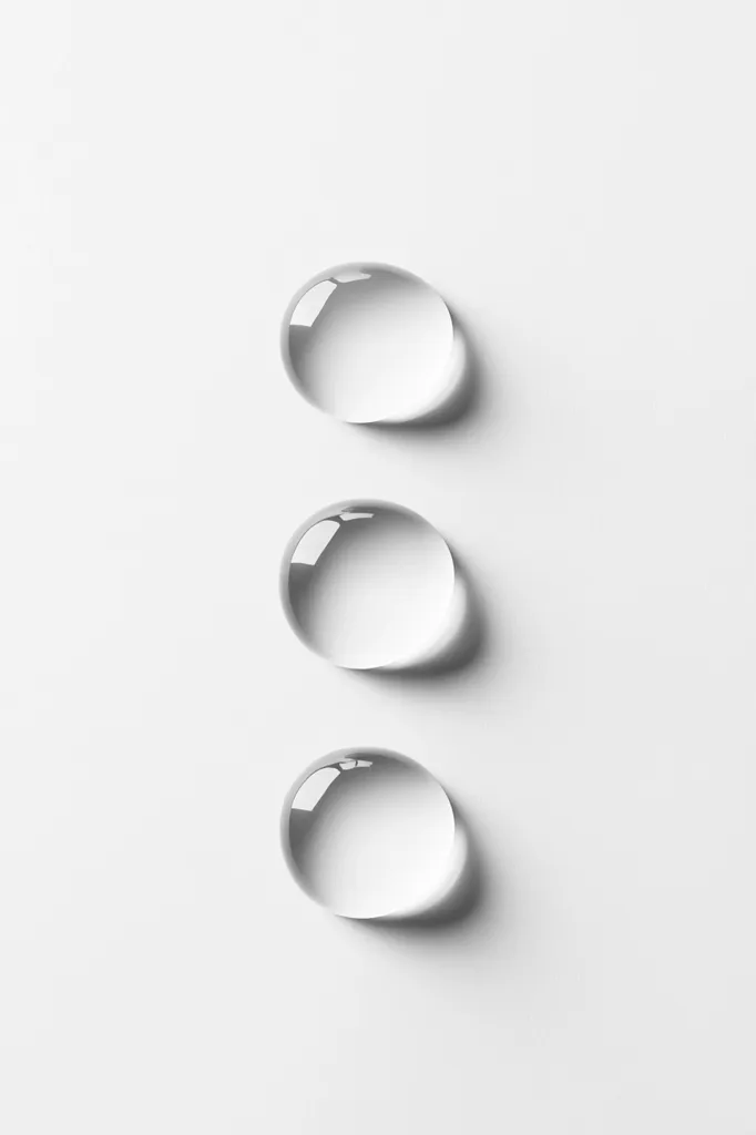 Three large, translucent water droplets are arranged vertically on a clean, white surface.  Each droplet is perfectly round, exhibiting a high degree of surface tension.  The droplets cast subtle shadows, enhancing their three-dimensional form against the minimalist backdrop.  The image is simple yet visually striking, emphasizing the purity and clarity of water.