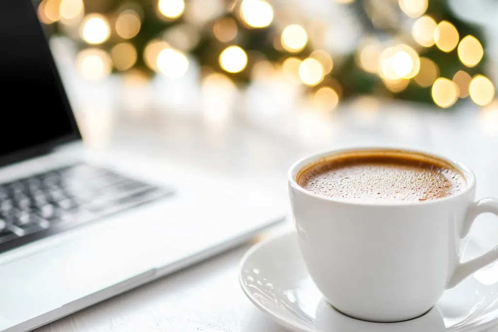 A cozy scene depicts a laptop partially visible on a white table, with a steaming cup of coffee in the foreground.  The background is softly blurred, featuring warm, golden bokeh lights suggesting a festive atmosphere. The overall image conveys a sense of warmth, comfort, and perhaps working from home during the holiday season.