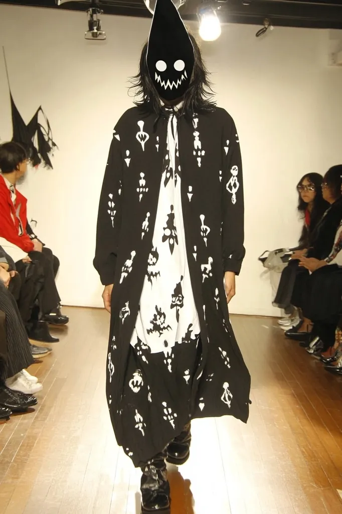 A model walks a runway wearing a long, black coat adorned with a white, abstract pattern.  The coat is worn over a matching black and white patterned shirt and pants. The model's face is concealed by a black, pointed mask with white eyes and a grinning mouth.  Other audience members are visible in the background, seated and observing the fashion show.  The overall aesthetic is dark and avant-garde.