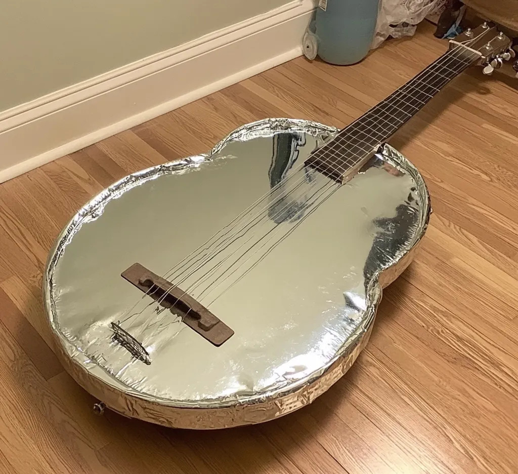 A guitar, uniquely modified, is showcased. Its body is entirely covered in shimmering aluminum foil, neatly wrapped and sealed, giving it a futuristic, almost otherworldly appearance. The wooden neck and fretboard contrast sharply with the metallic body. The instrument rests on a light brown hardwood floor, creating a striking visual juxtaposition. The overall impression is one of quirky ingenuity and artistic expression.