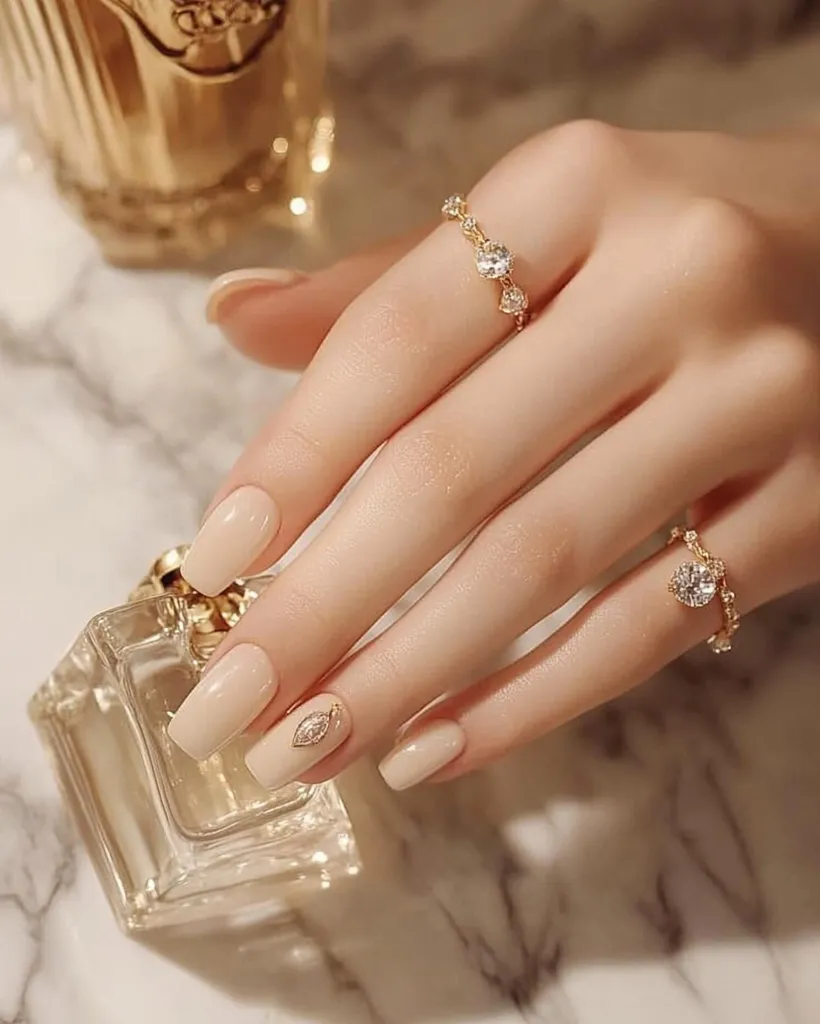 A close-up showcases elegantly manicured nails painted in a soft, neutral beige shade.  One nail features a delicate gold leaf accent.  The hand also displays two gold rings; one adorned with small, sparkling stones, and the other with a single, larger stone. The setting appears luxurious, with a partially visible gold perfume bottle in the background. The overall aesthetic is refined and minimalist.