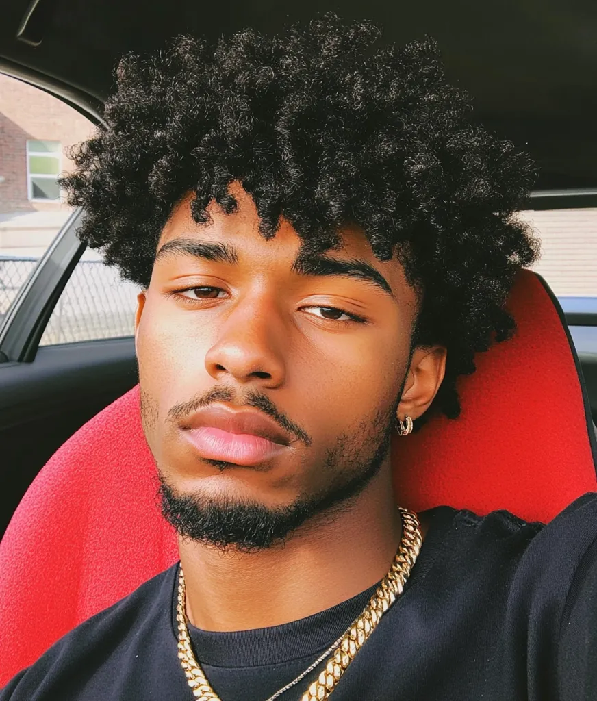 Close-up portrait of a young Black man with a full, curly afro. He has dark eyes, well-defined brows, and a goatee.  He's wearing a black shirt and a gold chain necklace, and small hoop earrings.  The background is partially visible, showing a red car seat and a glimpse of a building outside a car window.  His expression is serious and direct.