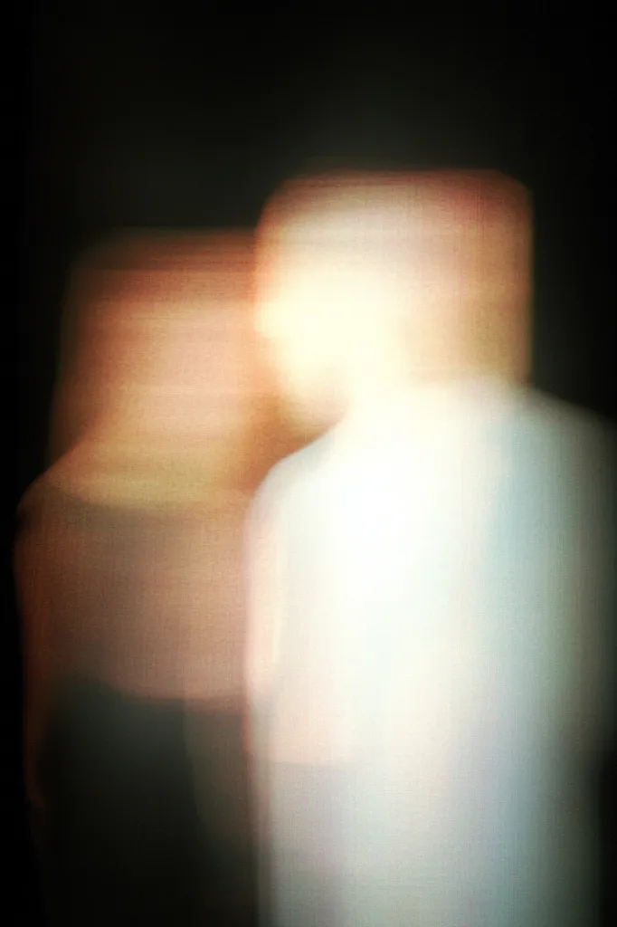 An abstract image depicts two blurred figures standing close together in a dimly lit space. The figures are indistinct, with soft, pastel colors predominating.  Their forms are suggestive of human shapes but lack sharp definition, creating a dreamy and ethereal atmosphere. The overall effect is one of mystery and intimacy.
