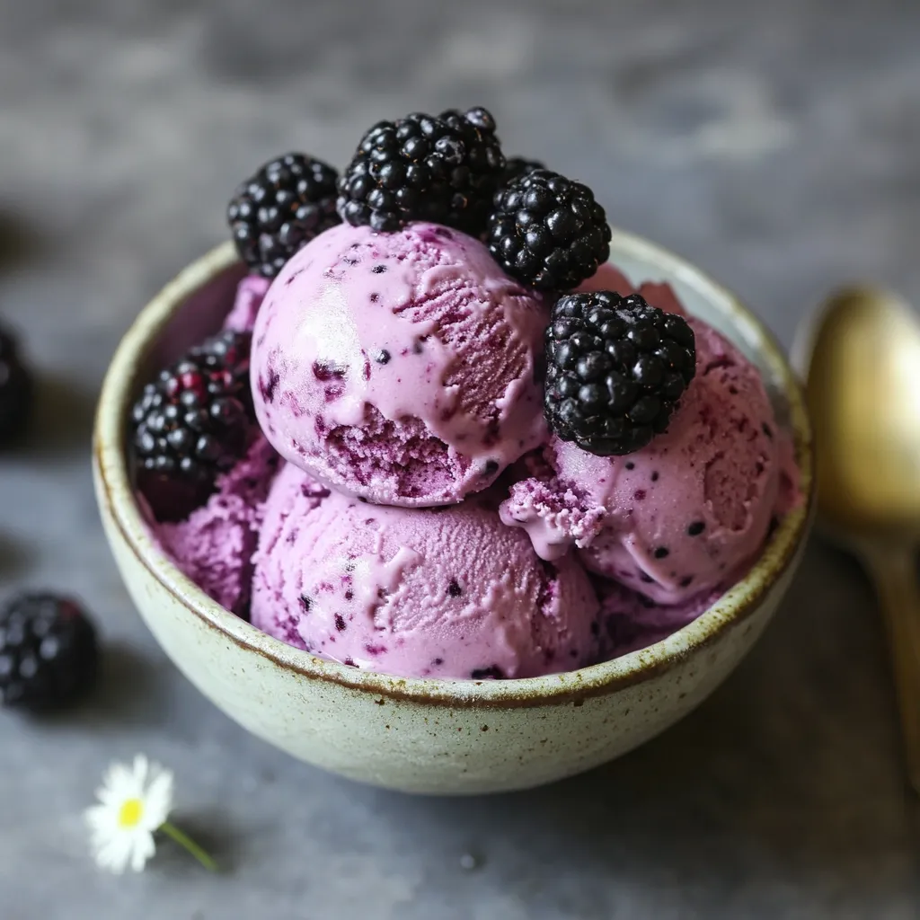 A bowl of homemade blackberry ice cream sits on a grey surface.  The pale purple ice cream is topped with fresh blackberries.  A few more blackberries are scattered nearby, along with a single daisy.  The image is styled with a shallow depth of field, focusing on the creamy texture and vibrant color of the dessert.  A gold spoon is partially visible in the background.