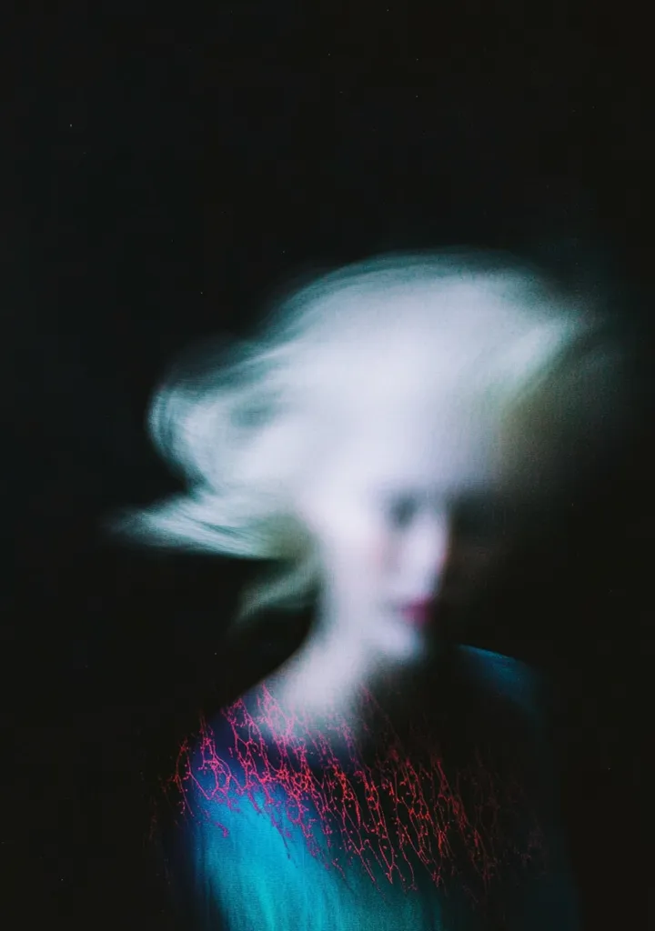 An abstract portrait of a woman with blonde hair, blurred and motion-like. The subject's features are indistinct, creating a dreamlike or ethereal quality.  She wears a teal top adorned with a vibrant pink, almost coral, design, contrasting with the muted background. The overall dark ambiance adds to the mysterious and artistic effect.