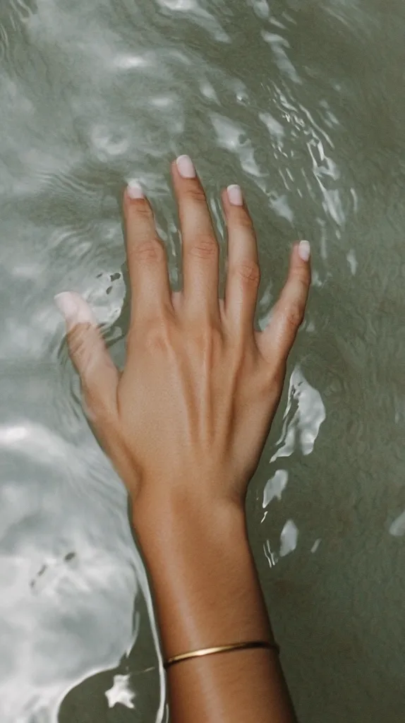 A woman's hand with neatly manicured nails rests gently on the surface of clear, still water.  The water's subtle ripples distort the view of the hand and arm, creating a serene and calming image. A delicate gold bracelet adorns her wrist.  The overall aesthetic is minimalist and emphasizes natural beauty and tranquility.