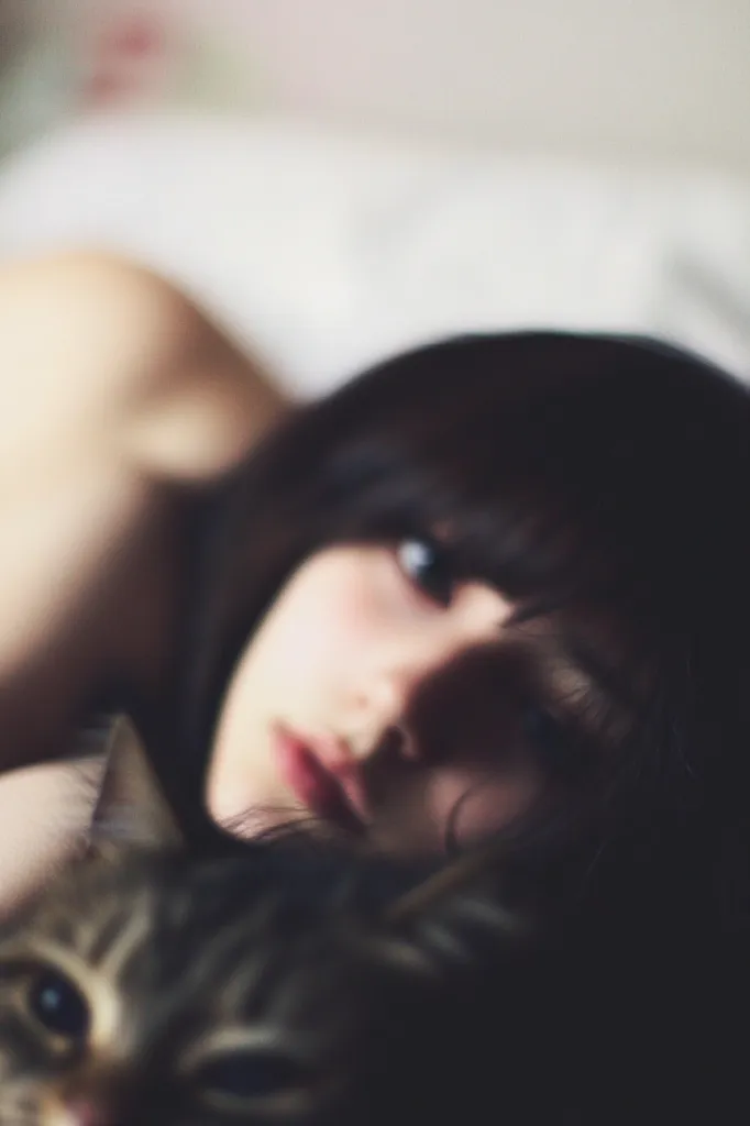 A young woman with dark hair lies on a bed, her face partially obscured by shadows.  Her eyes are visible, gazing slightly upward. A tabby cat rests near her face, its fur a blend of brown and gray. The overall mood is soft and intimate, with a slightly blurred focus emphasizing the closeness between the woman and her pet. The scene is dimly lit, contributing to the quiet and serene atmosphere.