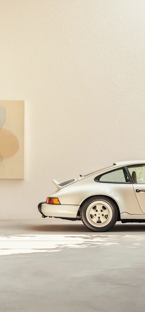 A classic white Porsche 911 Carrera is parked in a minimalist, light-filled garage.  The car's rear is prominently featured, showcasing its clean lines and design. A muted abstract painting hangs on the wall, complementing the car's understated elegance. The overall aesthetic is one of calm sophistication and modern simplicity.
