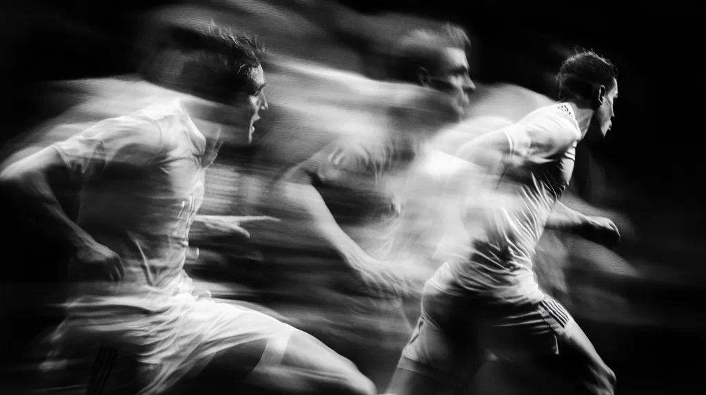 A black and white motion blur photograph captures the intense energy of a soccer player sprinting.  The image is heavily blurred, emphasizing speed and movement, with multiple superimposed figures suggesting rapid strides. The player is wearing a white jersey and shorts, their form dynamically captured in a moment of intense athleticism. The dark background isolates the figure, highlighting the dynamic energy of the run.