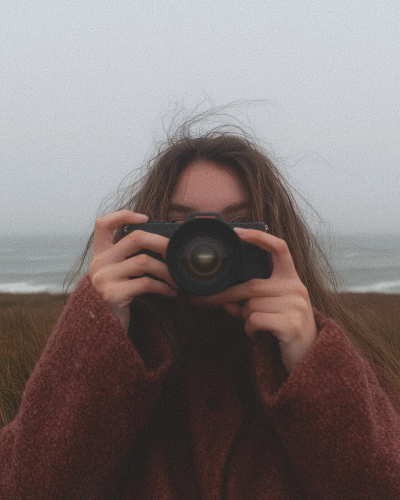 A person with long brown hair, wearing a maroon sweater, holds a camera to their face, obscuring their features.  The background is a blurred, overcast beach or coastal scene. The overall mood is muted and contemplative, suggesting a moment of quiet observation or photography. The focus is entirely on the act of taking a picture.