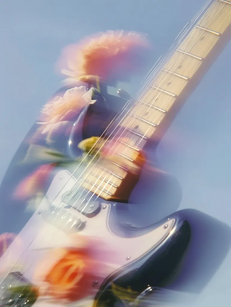 A blurry, dreamlike image shows an electric guitar with a light-colored body and dark-colored edges.  The guitar is partially obscured by a vibrant, out-of-focus overlay of peach and orange flowers, creating a surreal and artistic effect against a pale blue sky. The image suggests movement and energy.