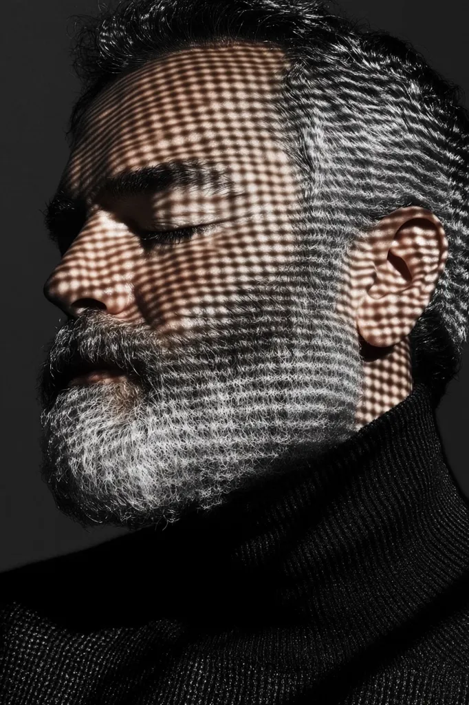 Close-up of a man's face, partially obscured by a grid-like shadow pattern.  He has graying hair and a salt-and-pepper beard. His eyes are closed, and he wears a dark turtleneck sweater. The image is predominantly black and white, creating a dramatic and moody atmosphere. The contrast between light and shadow is striking, emphasizing the textures of his skin and beard.