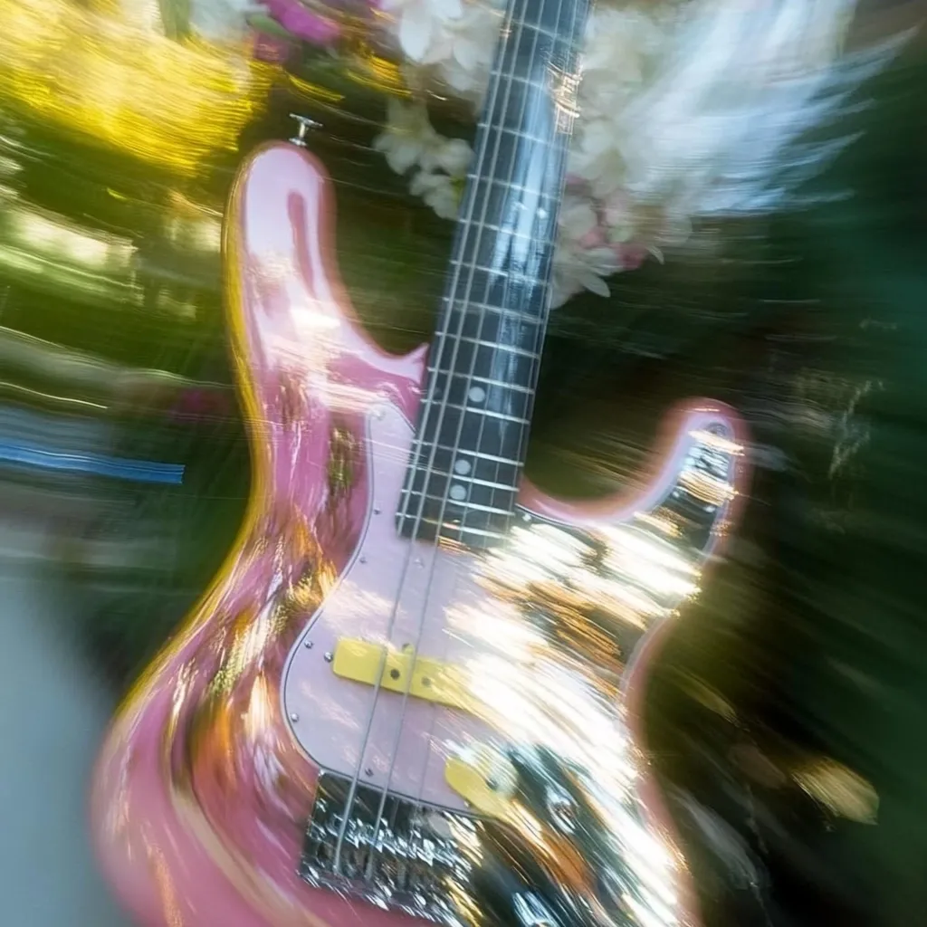 A pink bass guitar is shown in a blurred, motion-filled image.  The instrument is positioned slightly angled, with the body reflecting light.  The background is a vibrant mix of blurred greens and yellows, suggesting an outdoor setting.  The overall effect is dreamy and ethereal, emphasizing the vibrant color of the guitar.