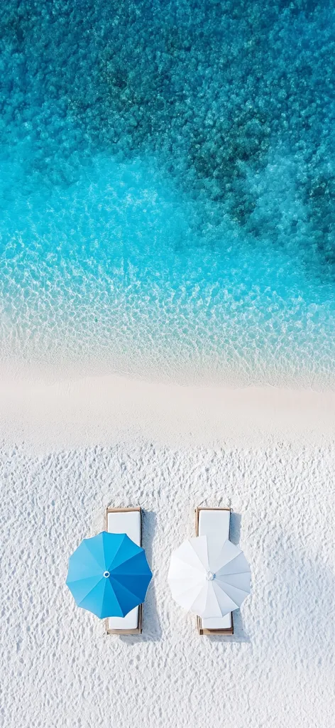 An aerial view of a pristine beach with two lounge chairs and parasols.  One parasol is bright blue, the other white.  The sand is white and powdery, meeting the turquoise ocean water in a gentle gradient.  The scene is peaceful and idyllic, suggesting a luxurious tropical getaway.