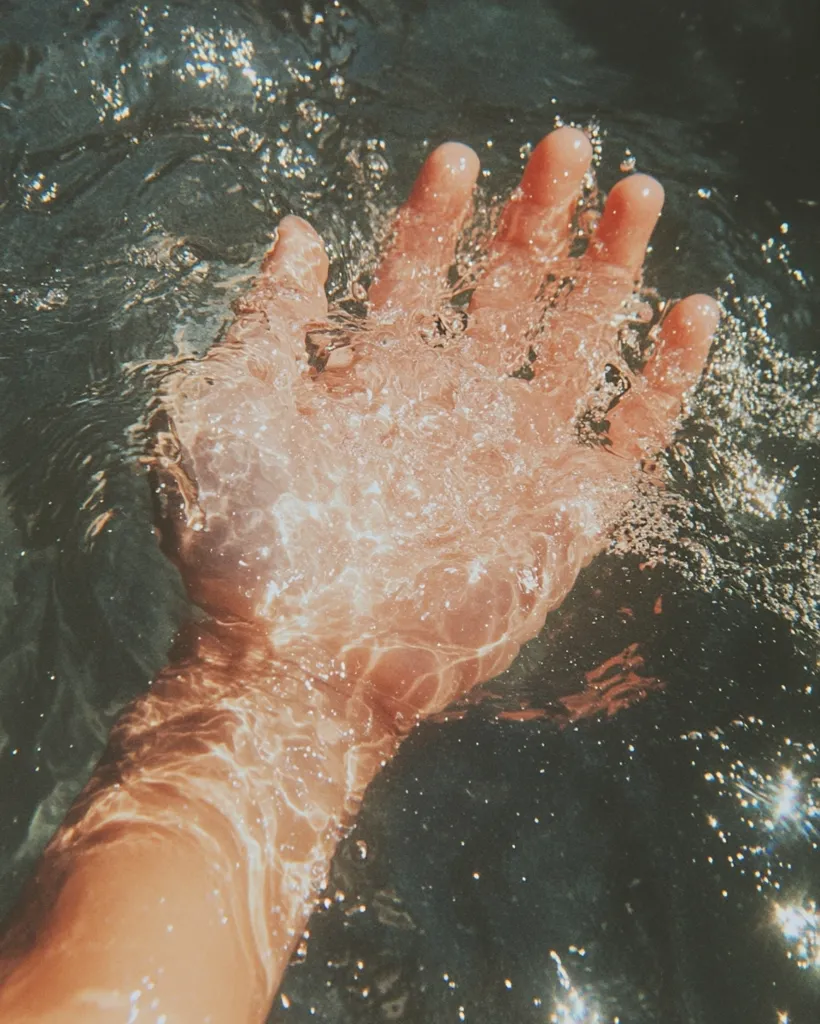 A hand emerges from clear, sunlit water, its fingers gently parting the surface.  Bubbles cling to the skin, creating a shimmering effect. The water is dark below, contrasting with the bright, almost ethereal quality of the hand and the light above. The image evokes a sense of calm and refreshing coolness.